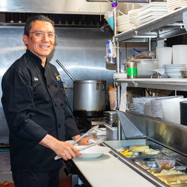 A smiling Bella Pasta chef in a black uniform prepares food in a busy stainless steel kitchen.